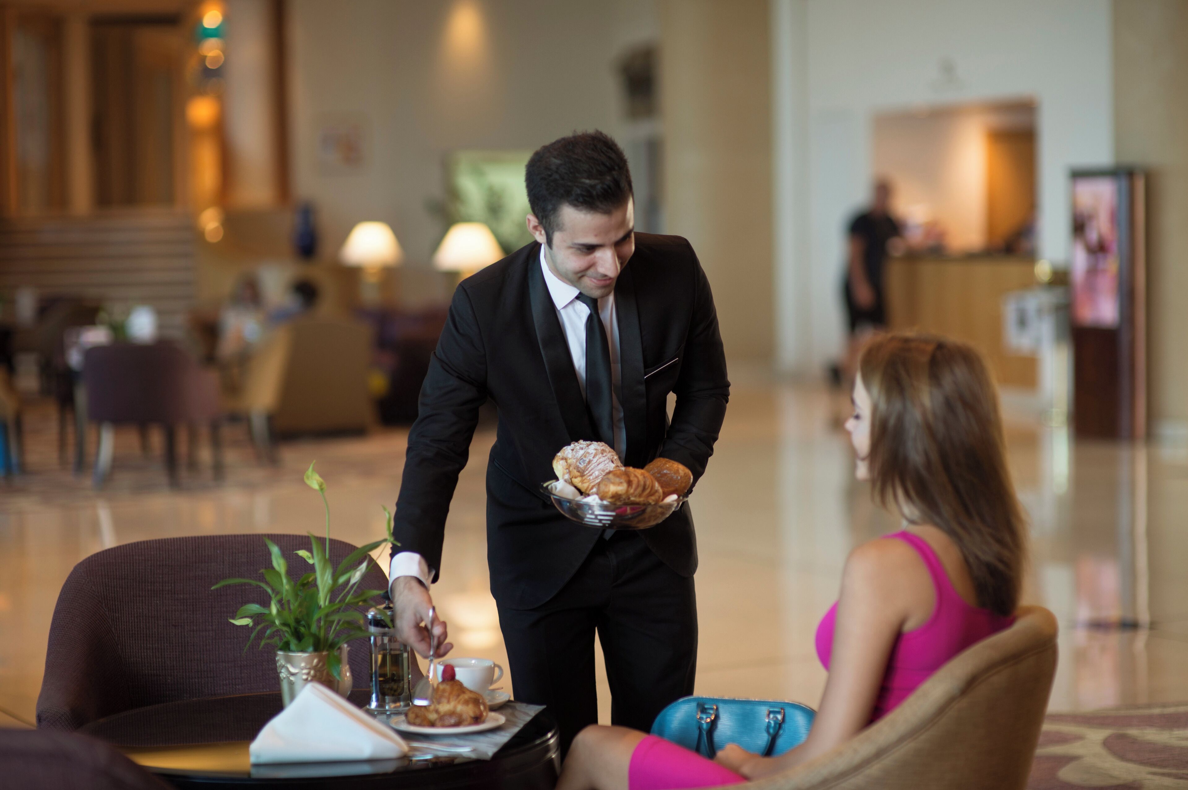 Un homme en costume noir sert de la nourriture à une femme en robe rose dans le hall d’un hôtel, où se trouvent un comptoir d’accueil et des chaises.