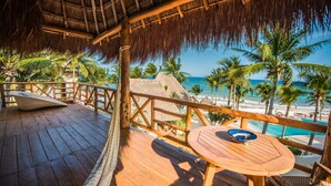 Une terrasse en bois, surmontée d’un toit de chaume, qui offre une vue sur une plage bordée de palmiers et sur l’océan.