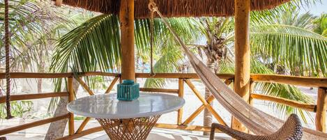 Zona de estar al aire libre con techo de paja, mobiliario de mimbre y una mesa de vidrio, con vistas a una playa con palmeras.