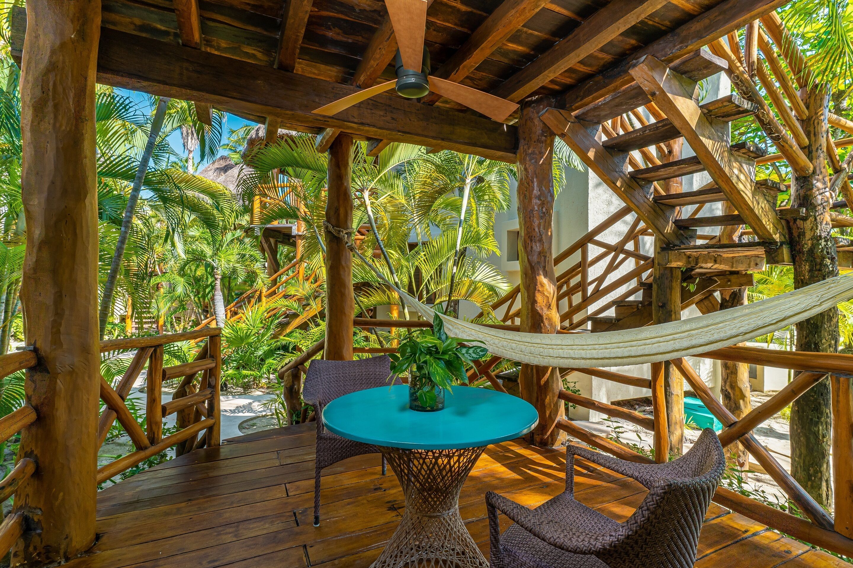 A wooden deck with a hammock, a round table, and chairs, surrounded by lush tropical vegetation.