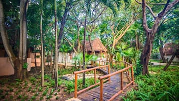 A wooden bridge leading to a thatched-roof structure surrounded by dense tropical foliage.