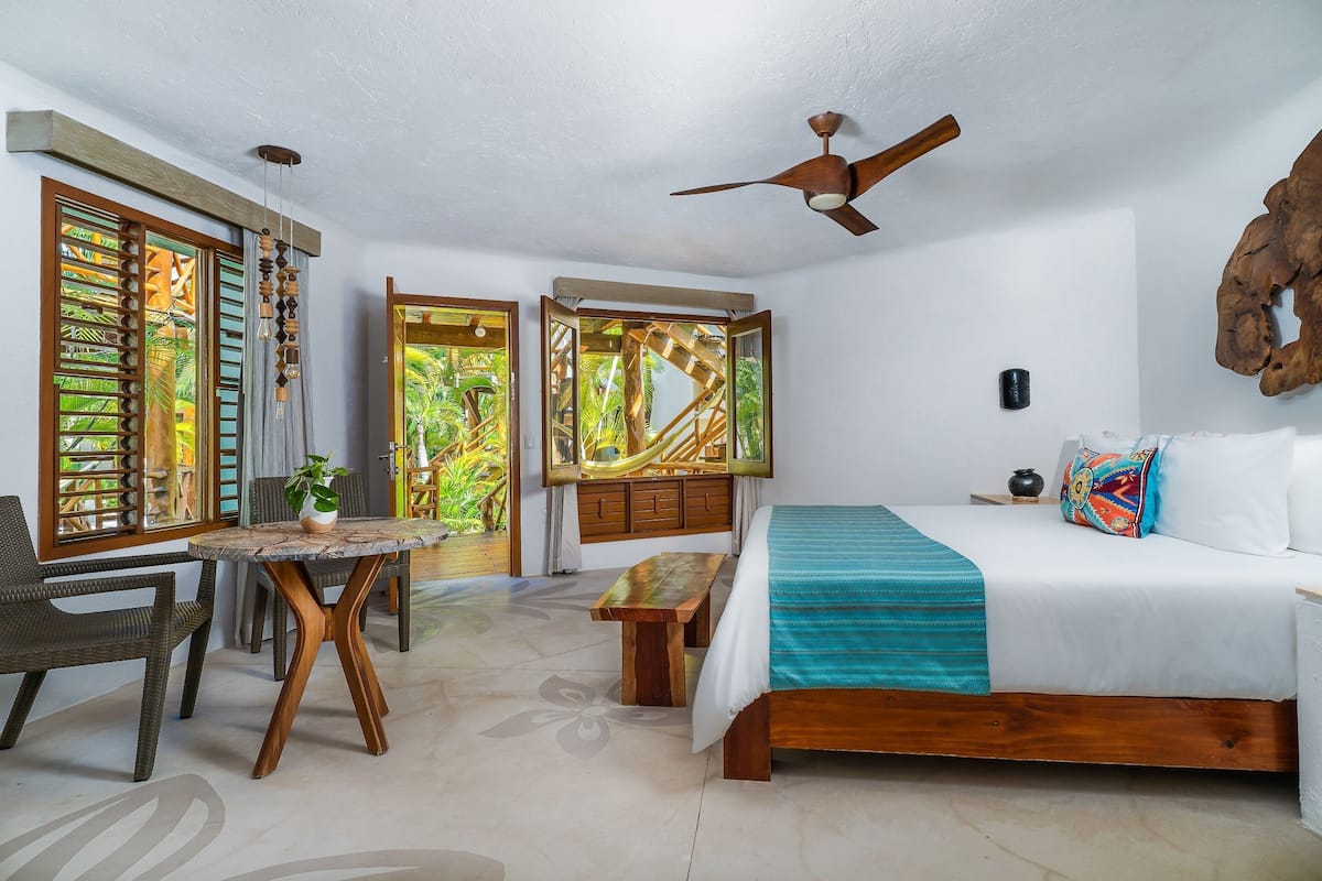 A bedroom with a bed, a desk, and a chair. There are windows with wooden shutters and a ceiling fan.