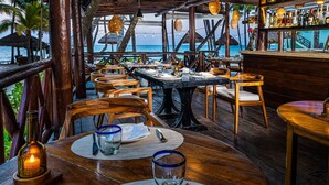 Un restaurant en plein air avec des tables et des chaises en bois, un bar avec des bouteilles exposées et une vue sur l’océan.