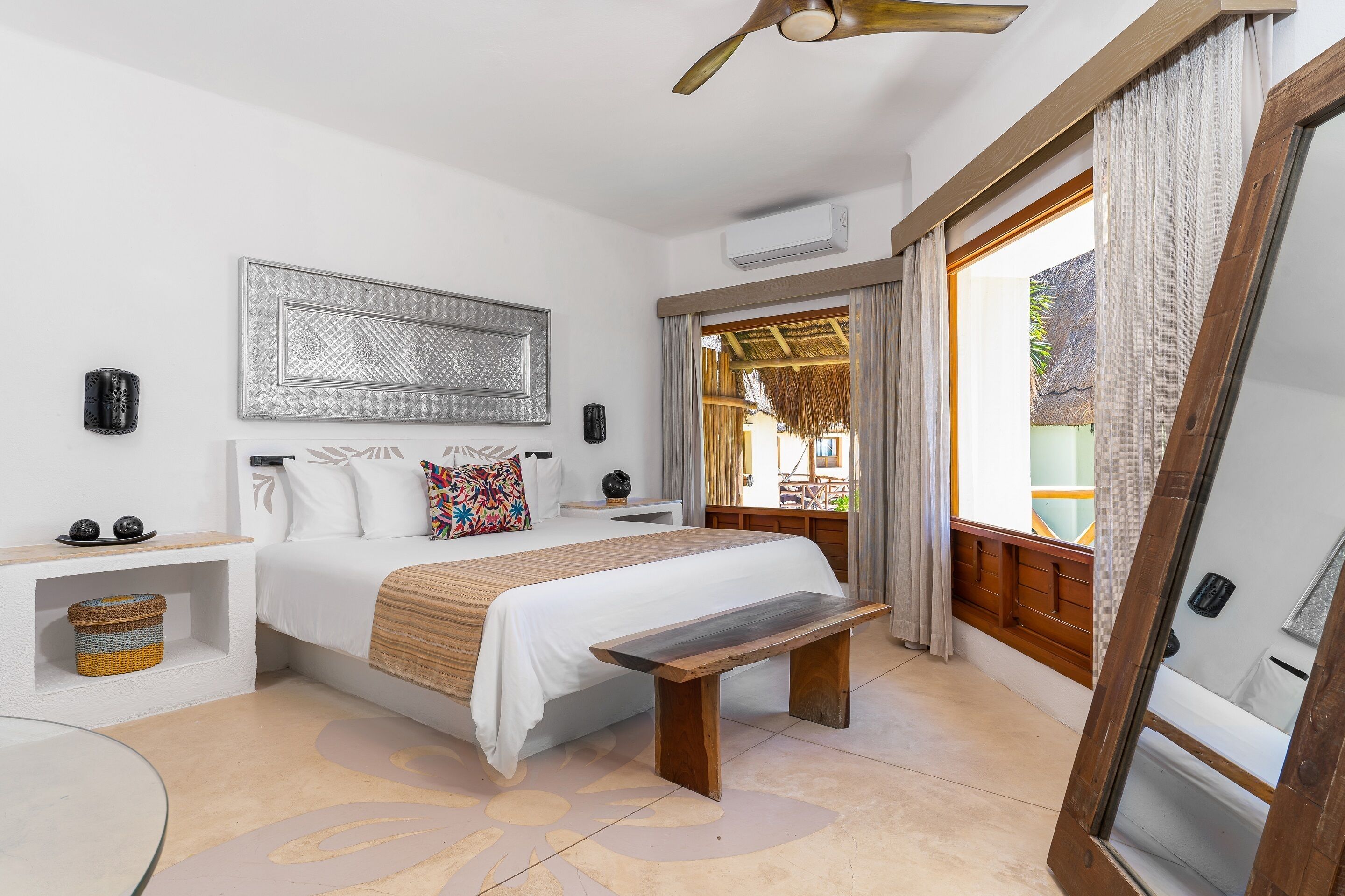 A bedroom with a bed, bedside tables, a bench, a ceiling fan, and a large window with curtains.