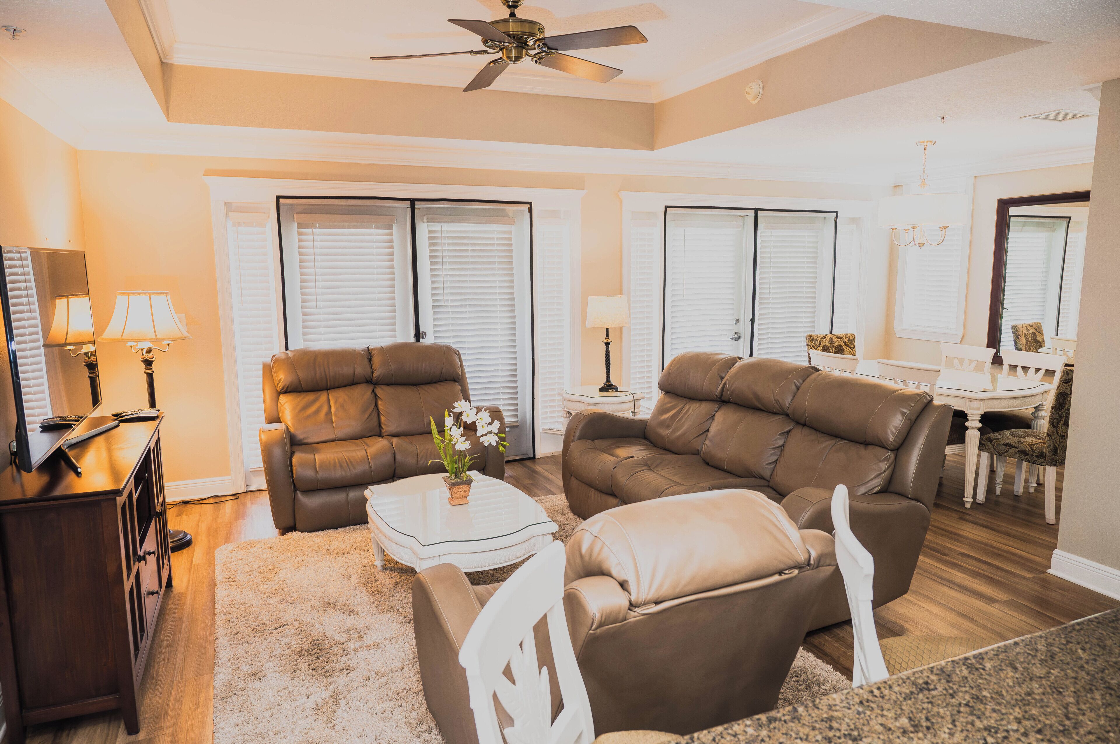 A living room with a brown leather sofa, a white coffee table, and a TV on a wooden stand. There are two lamps on side tables, and a ceiling fan is visible.