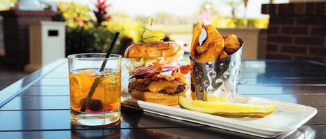 Comida que incluye una hamburguesa, papas fritas y una bebida sobre una mesa con vista a palmeras.