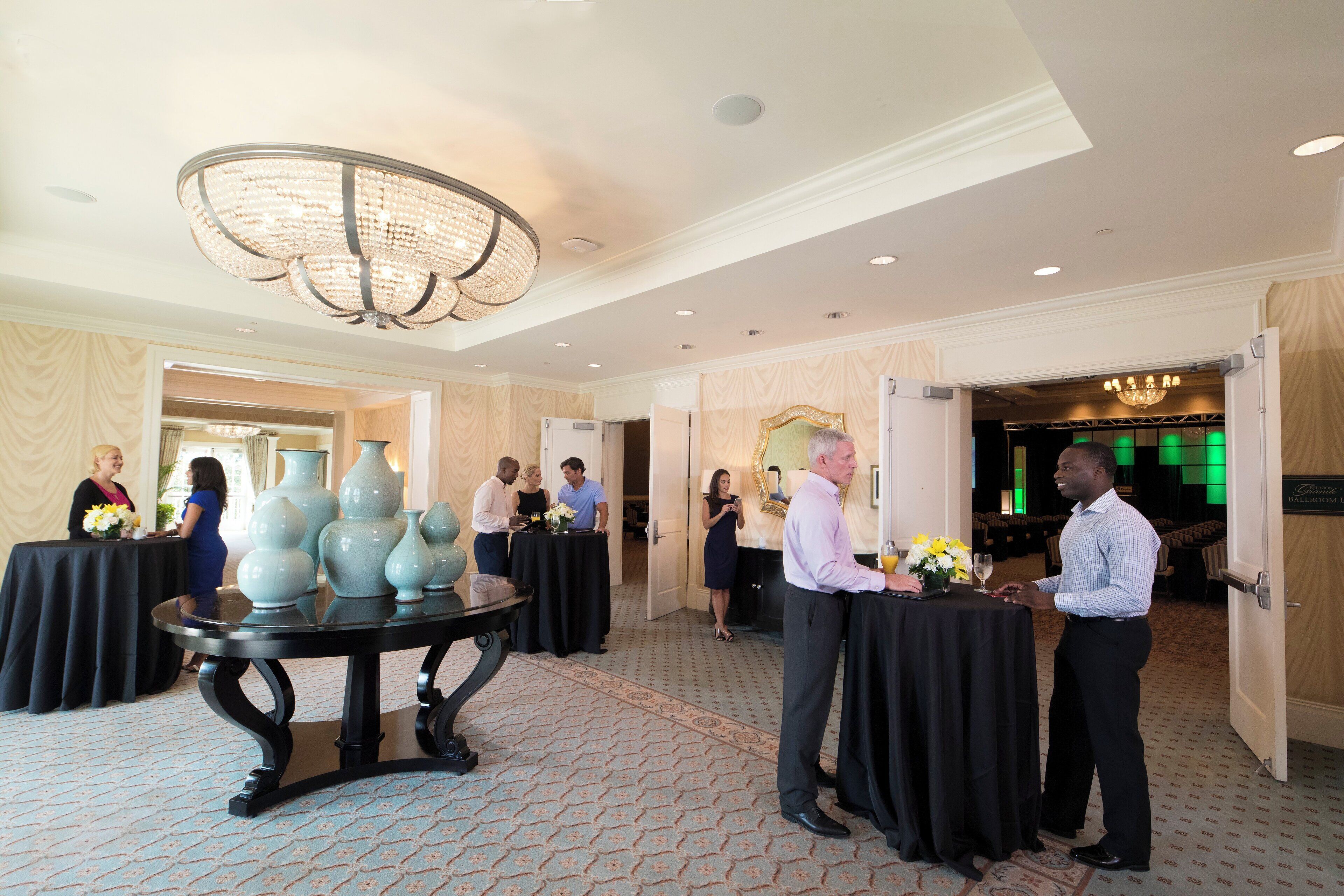 A hotel lobby with a chandelier, tables with floral centerpieces, and a patterned carpet.