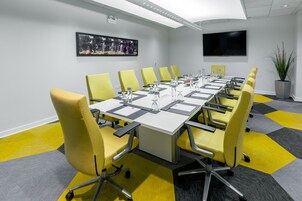 A conference room with a long table, yellow chairs, a flat-screen TV, and a potted plant.