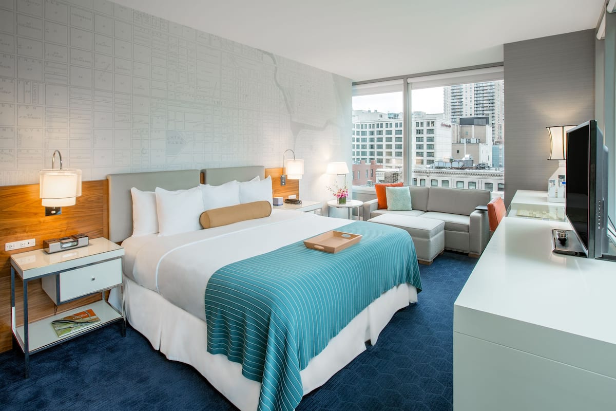 A modern hotel room with a large bed, a desk, a chair, and a view of the city.
