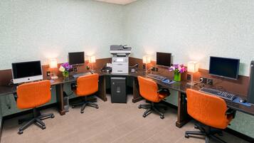 Una sala de conferencias con cuatro sillas de oficina color naranja, una mesa central con una impresora, dos monitores y dos lámparas.