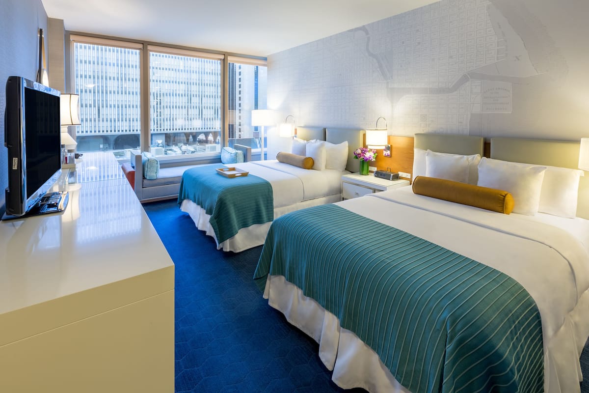 A hotel room with two beds, a TV, a desk, and a large window with a city view.
