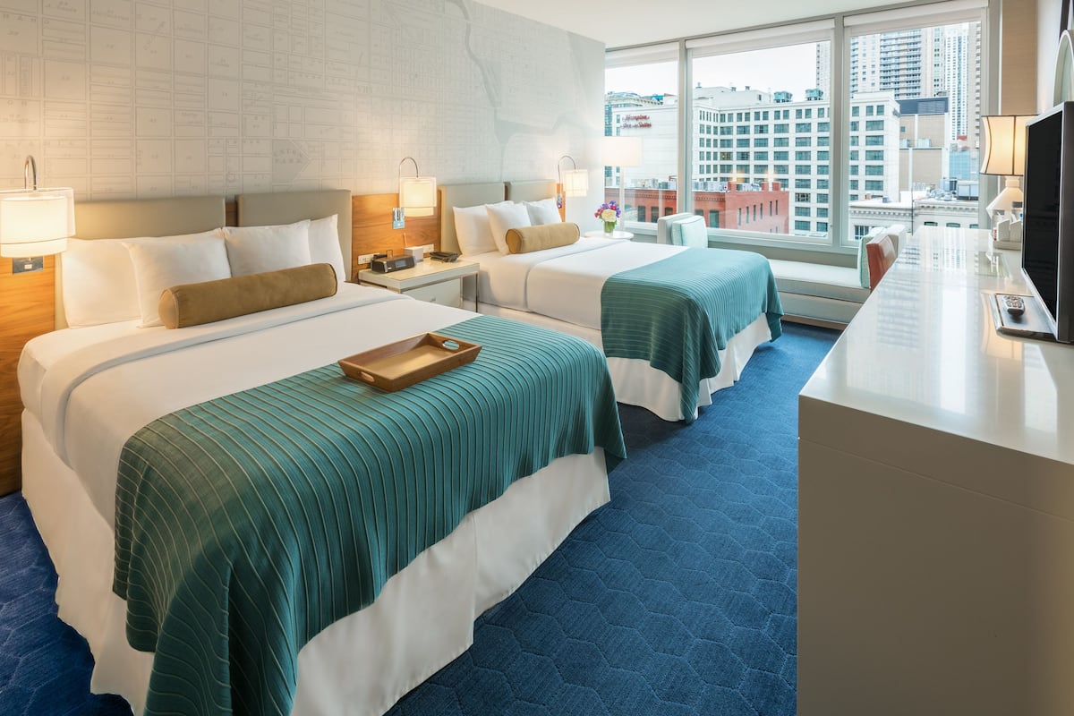 A hotel room with two beds, a desk, a TV, and a large window with a city view.