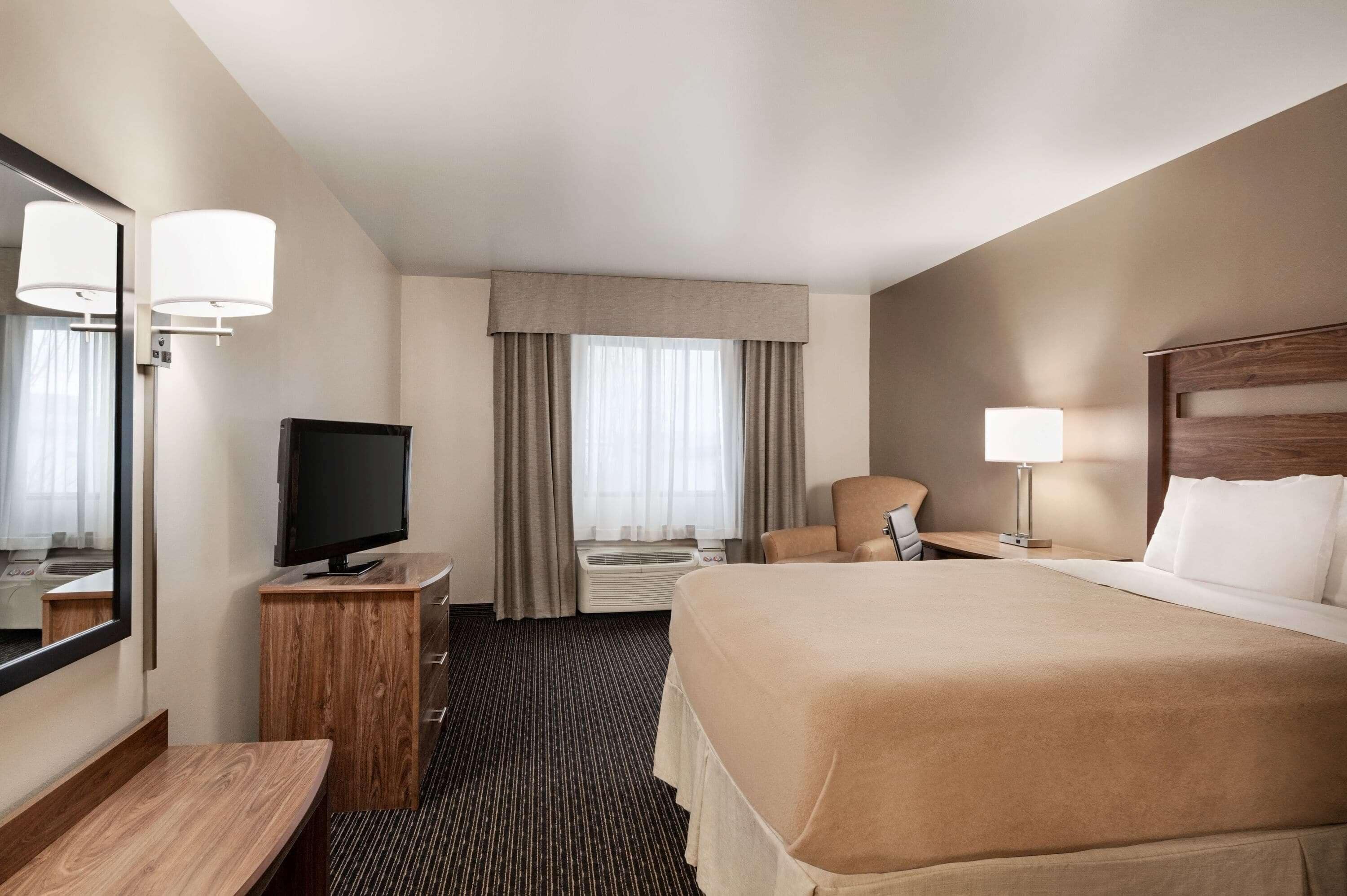 A hotel room with a large bed, a desk with a television, a chair, a bedside table with a lamp, and a large window with curtains.