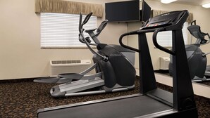 A fitness room with treadmills and a stationary bike, a television on the wall, and a window with blinds.