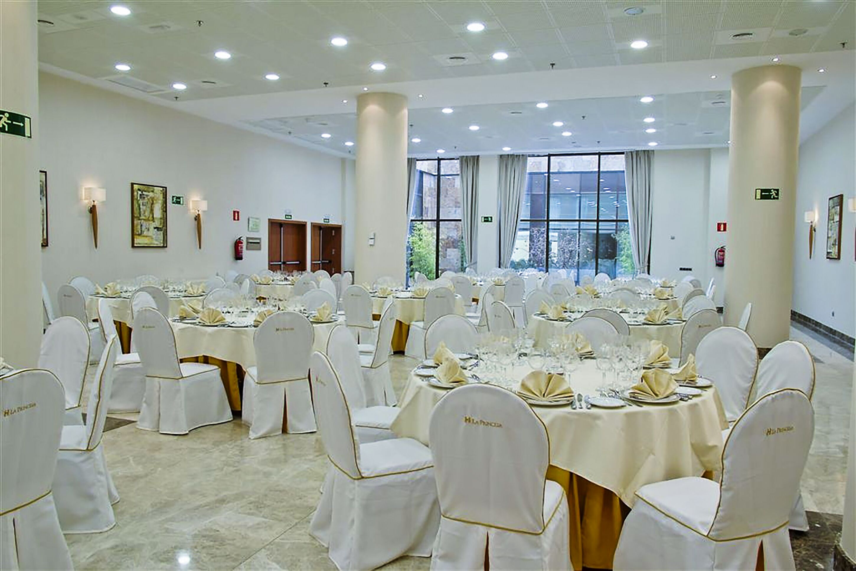 Un amplio salón de banquetes con mesas redondas dispuestas para comer, sillas blancas y grandes ventanales que dejan entrar luz natural.