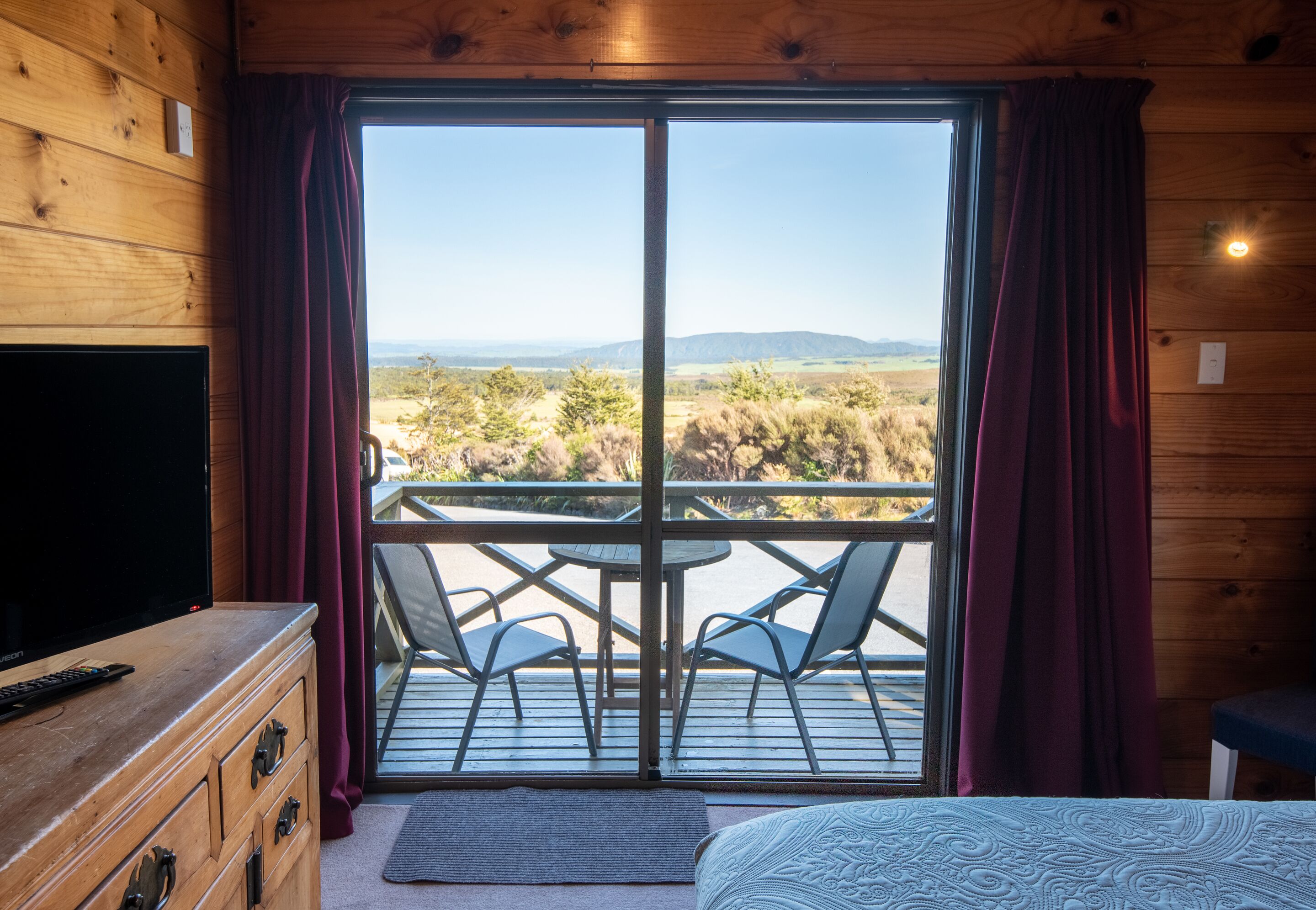 A room with a wooden interior, a TV on a dresser, a bed, a balcony with a table and chairs, and a view of the countryside.