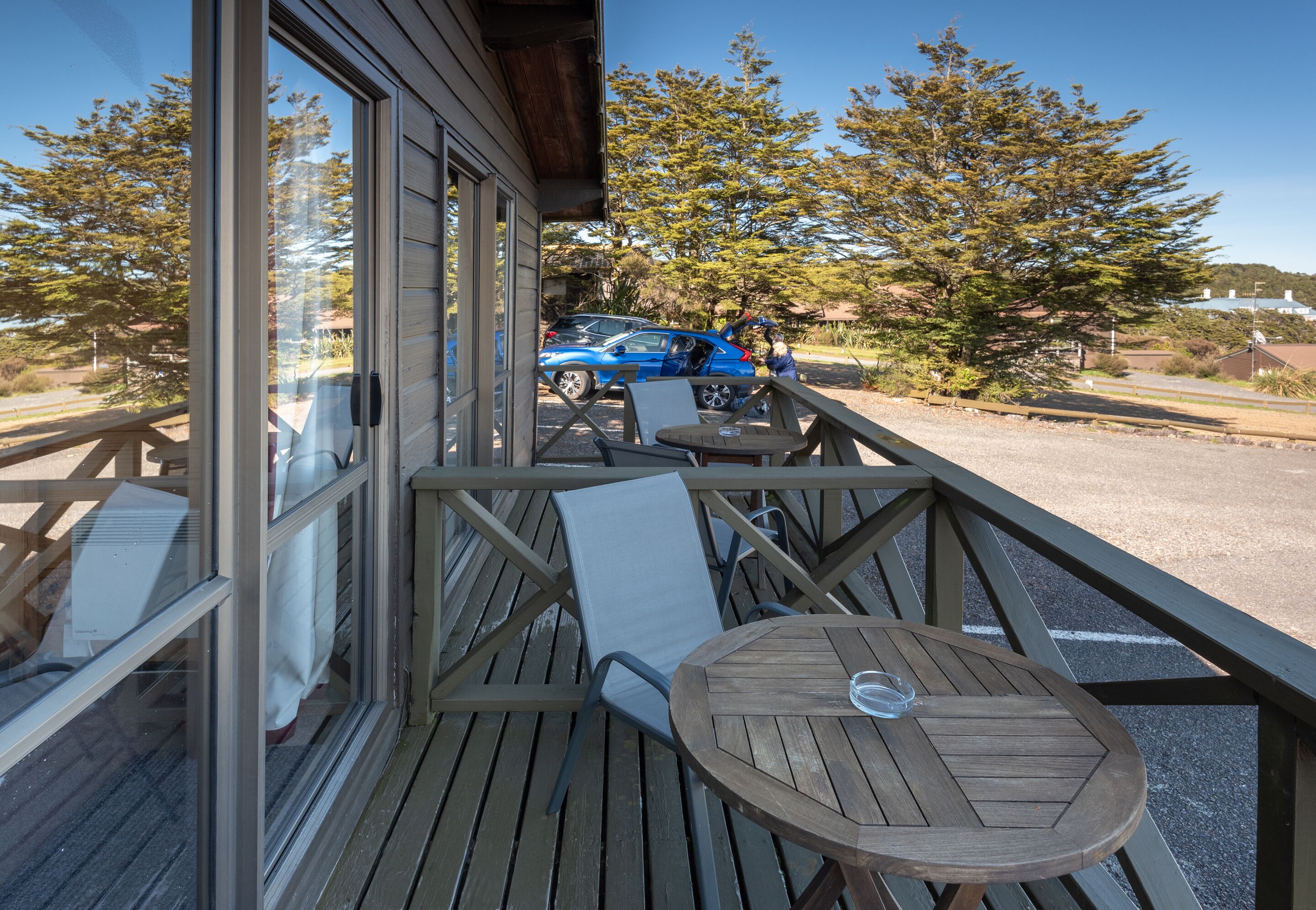 Une terrasse en bois avec une table et des chaises, donnant sur un parking où se trouve une voiture bleue.