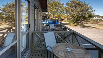 Une terrasse en bois avec une table et des chaises, donnant sur un parking où se trouve une voiture bleue.