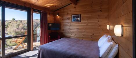 A wooden cabin room with a bed, a chair, a table, and a large window offering a view of the outdoors.