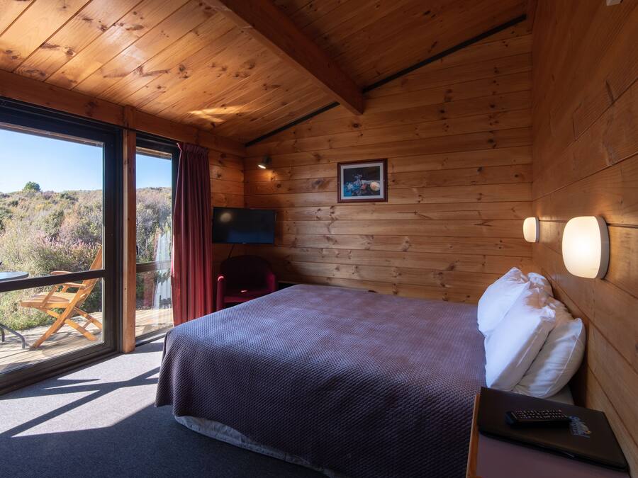 Quarto de cabana de madeira com cama, cadeira, mesa e uma grande janela com vista para o exterior.