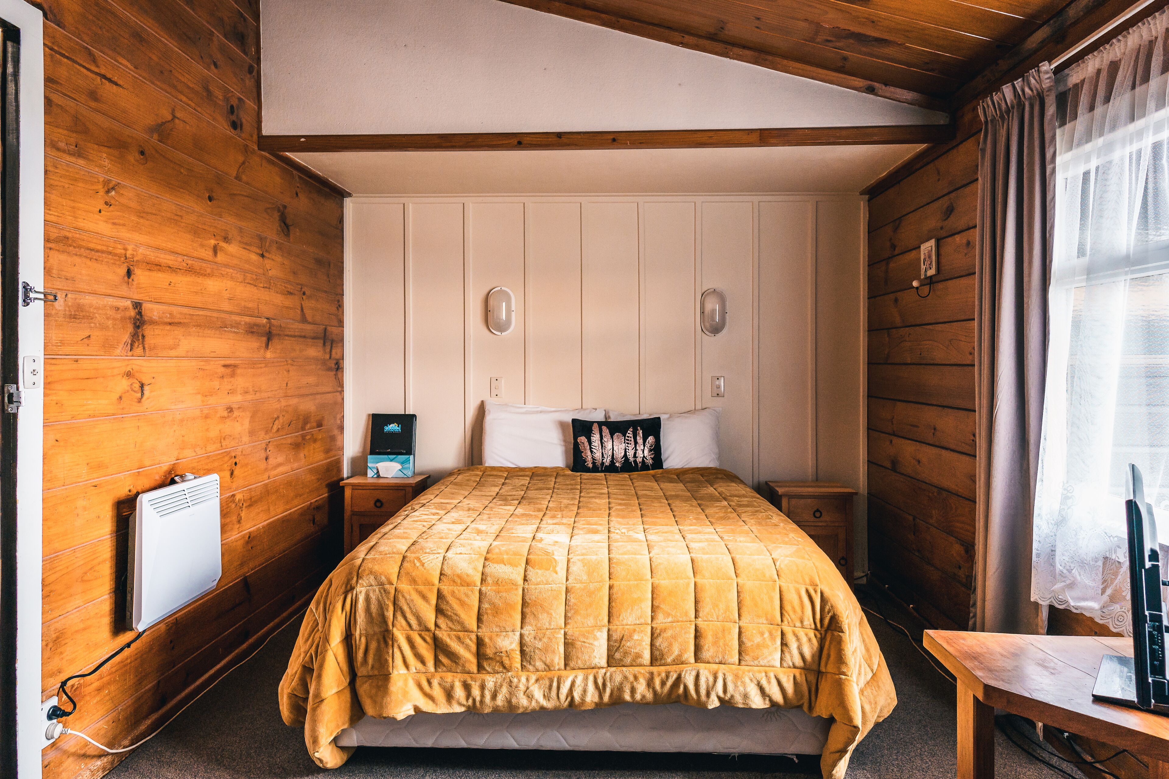 A room with a bed, wooden walls, a small bedside table, a nightstand, a heater, and a window with curtains.