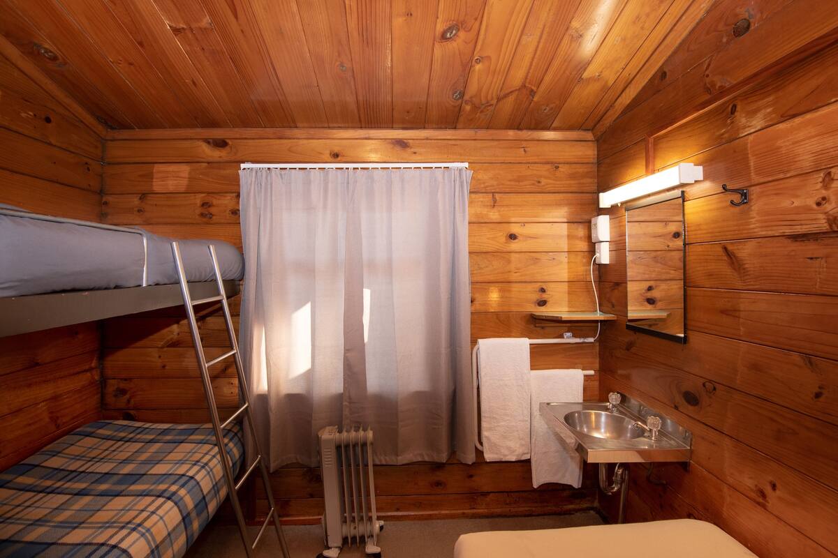 Eine Hütte mit einem Etagenbett, einem kleinen Badezimmer inklusive Waschbecken und Spiegel sowie einem Fenster mit Vorhängen.