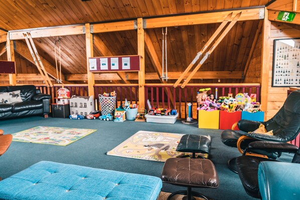Une pièce chaleureuse avec un plafond haut, des poutres apparentes, un canapé en cuir noir, un pouf bleu, et une variété de jouets et de jeux pour enfants.