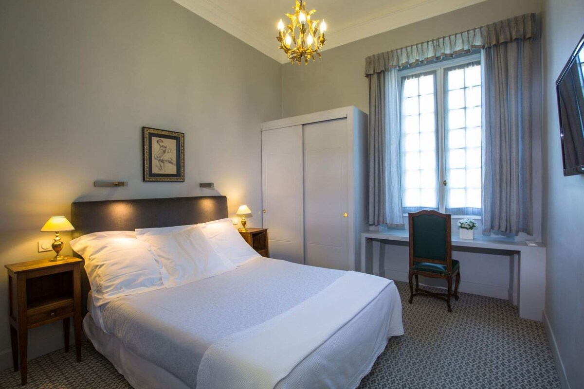 A hotel room with a bed, two bedside lamps, a chair, a window with curtains, a wall-mounted TV, and a chandelier.