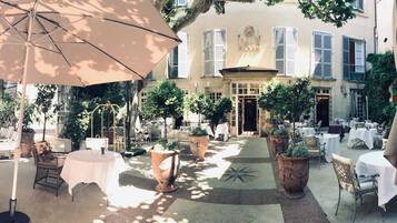 An outdoor patio with tables and chairs, surrounded by potted plants and a large umbrella.