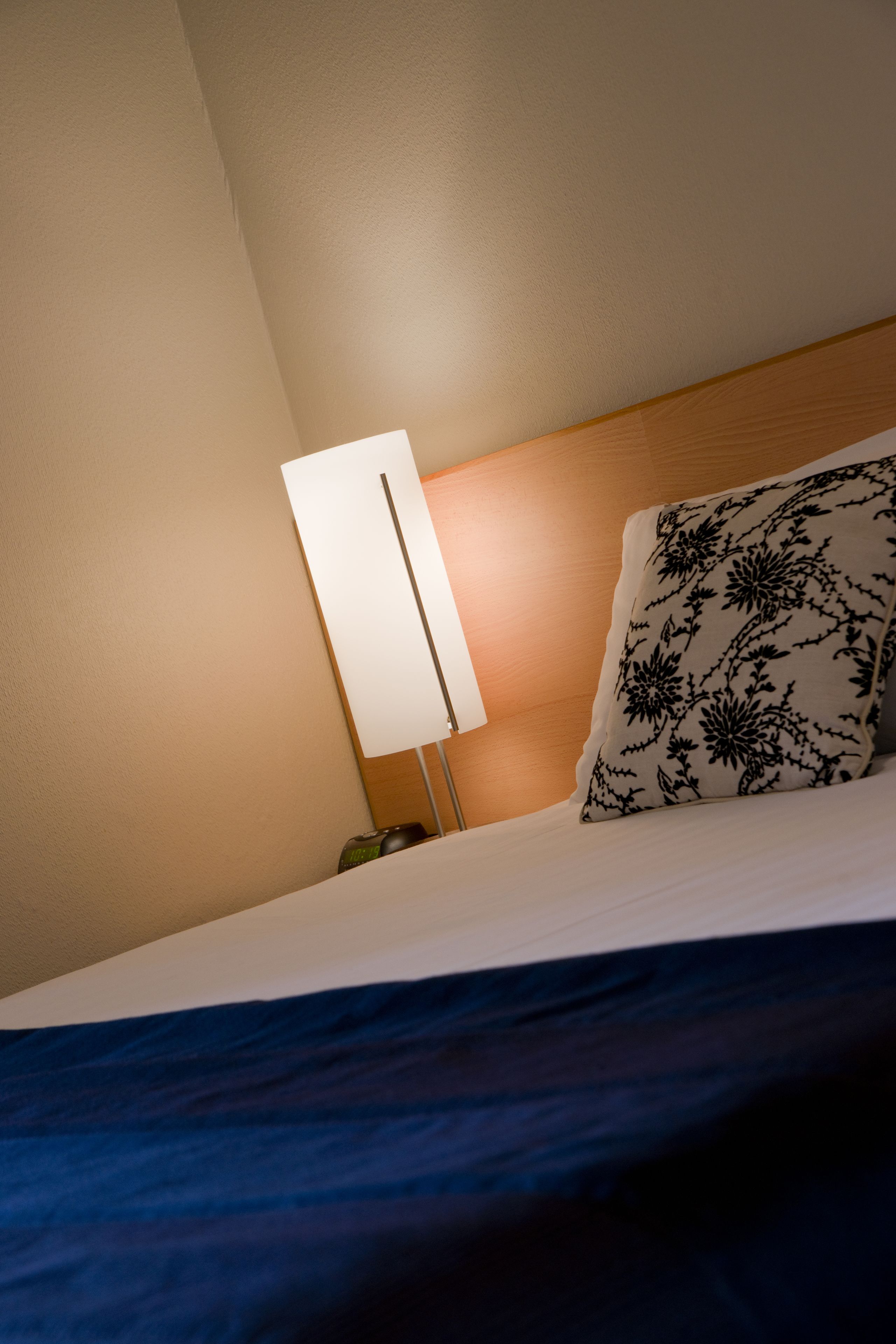 A neatly made bed with a blue bedspread, a white pillow with a black floral pattern, and a bedside lamp with a white shade.