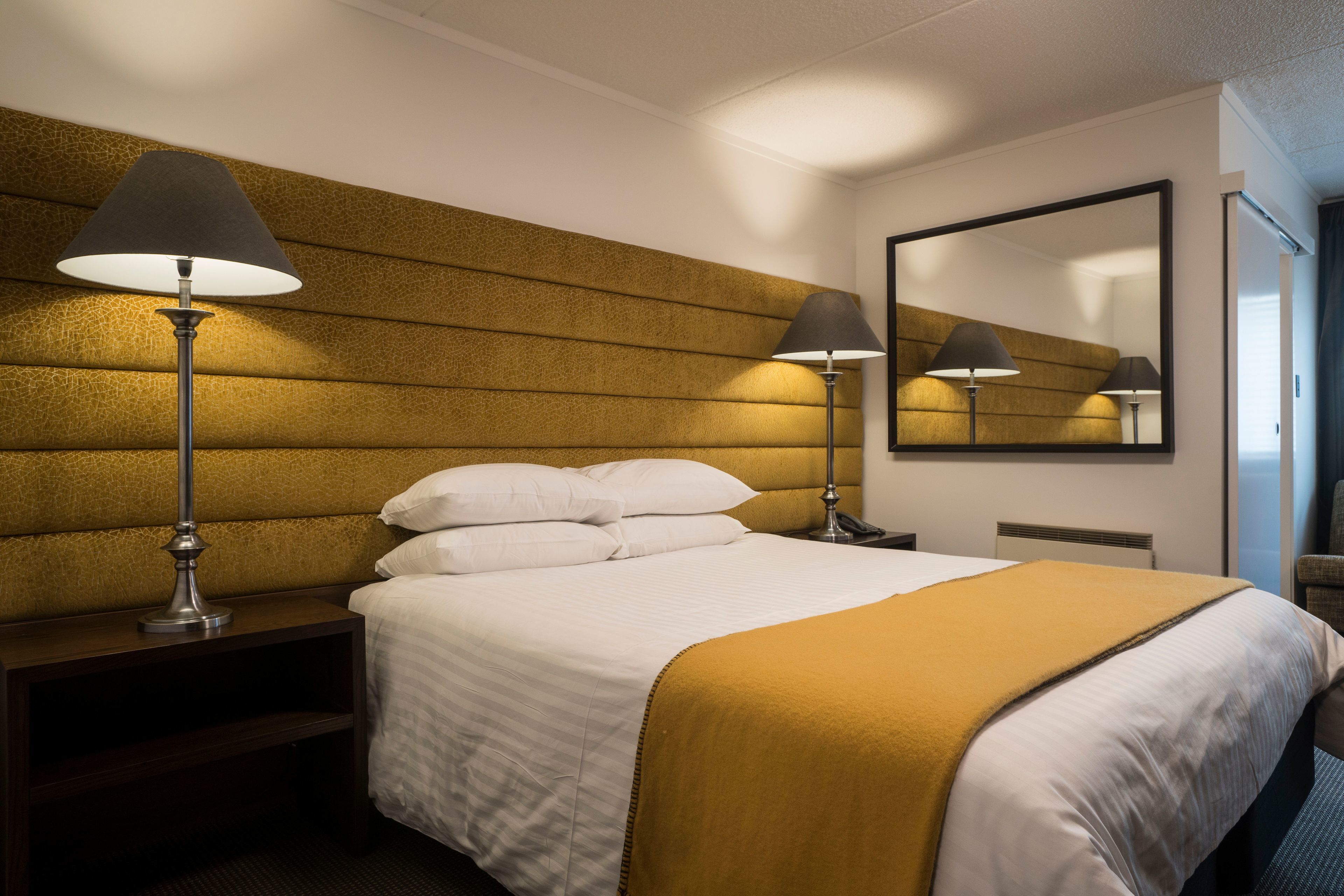 A hotel room with a large bed, two bedside lamps, a headboard with a textured pattern, a mirror on the wall, and a nightstand.