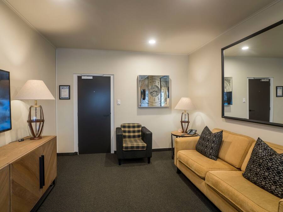 Quarto de hotel com sofá, uma cadeira, escrivaninha com luminária, espelho grande e relógio de parede.