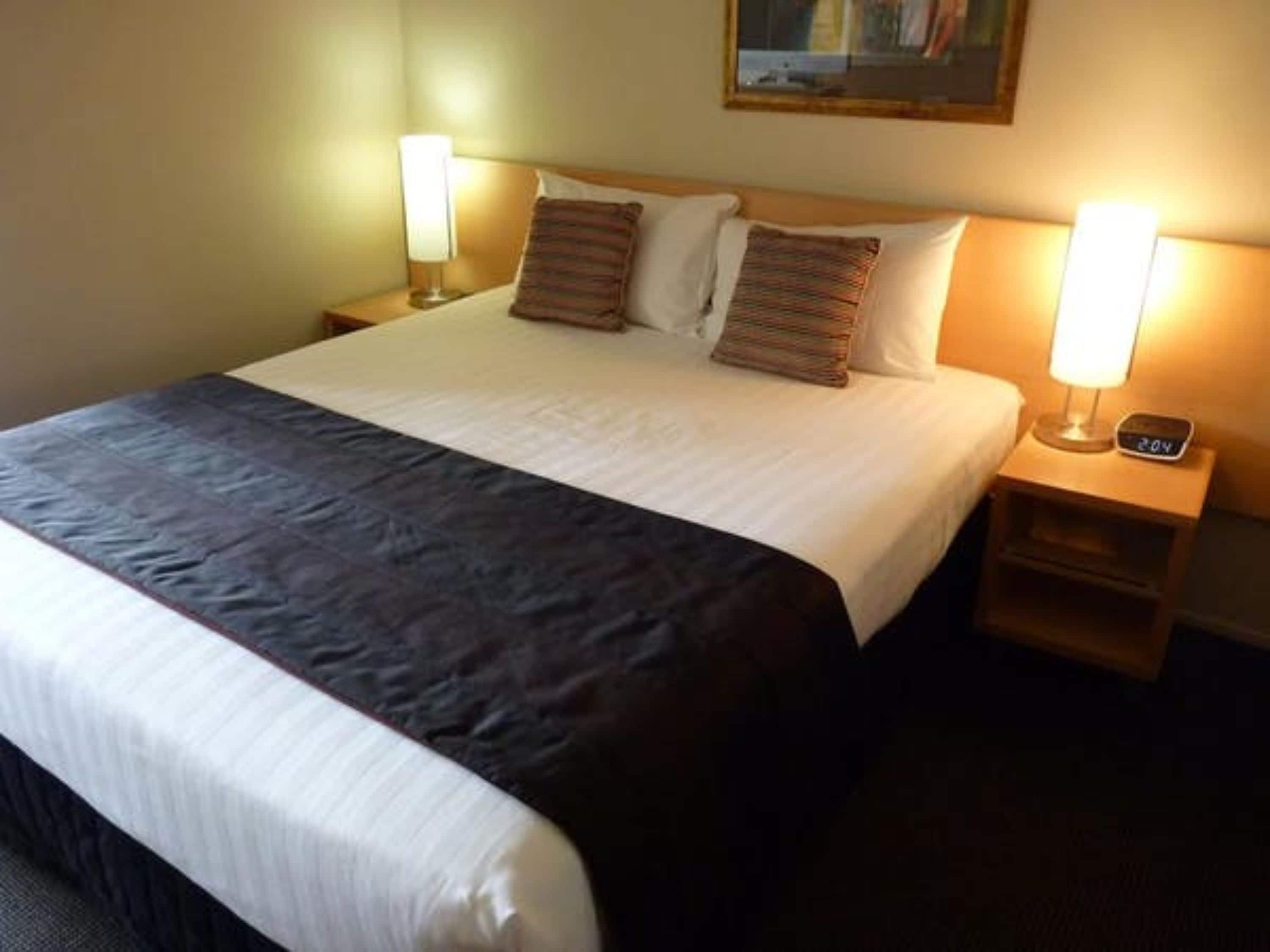 A neatly made bed with a dark-colored bedspread and two pillows, a bedside table with a lamp, and a clock displaying the time.