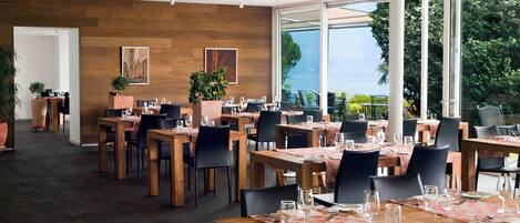 A spacious dining area with wooden tables and chairs, large windows offering a view of the outdoors, and potted plants adding greenery.