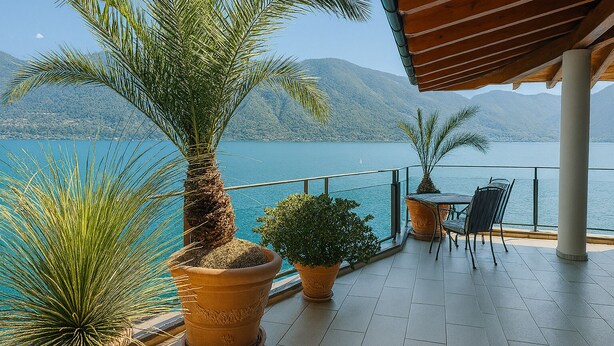 A terrace with potted plants, a table with chairs, and a view of a lake and mountains. - Hotel Collinetta (Ascona)