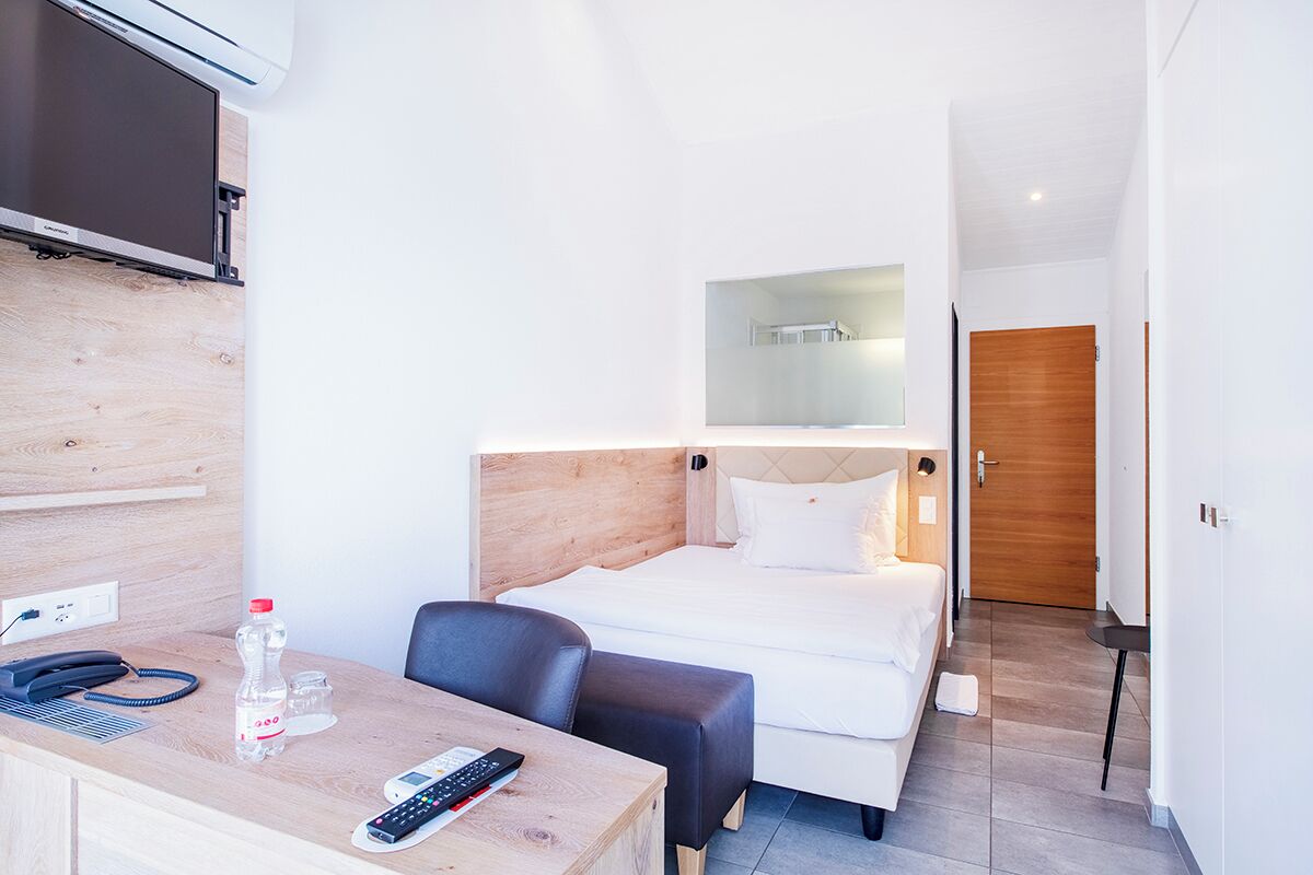 Ein Hotelzimmer mit Bett, Schreibtisch mit Telefon und Fernbedienung, Stuhl, Holz-Kopfteil und Spiegel über dem Bett.
