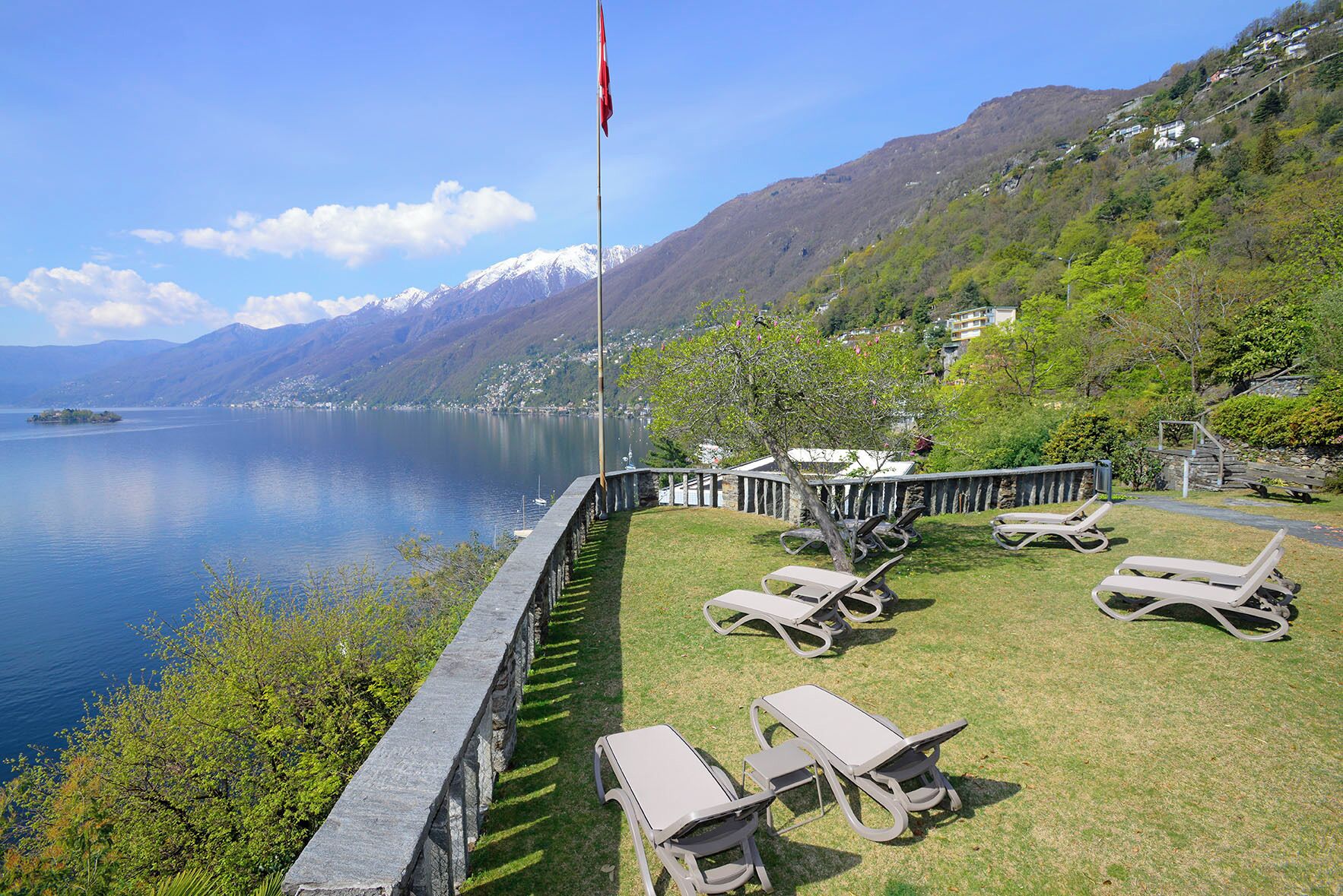 Terraza junto al lago con sillones de descanso, una bandera y un muro de piedra con vistas a una montaña y un lago.