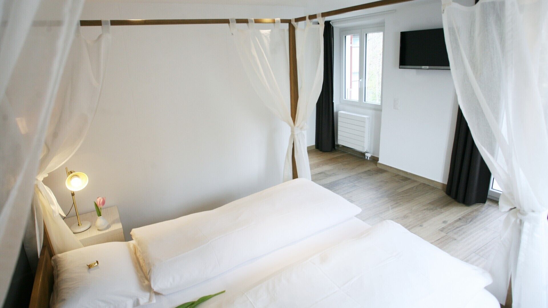 A bedroom with a bed, a bedside table with a lamp, a window, and a TV on the wall.