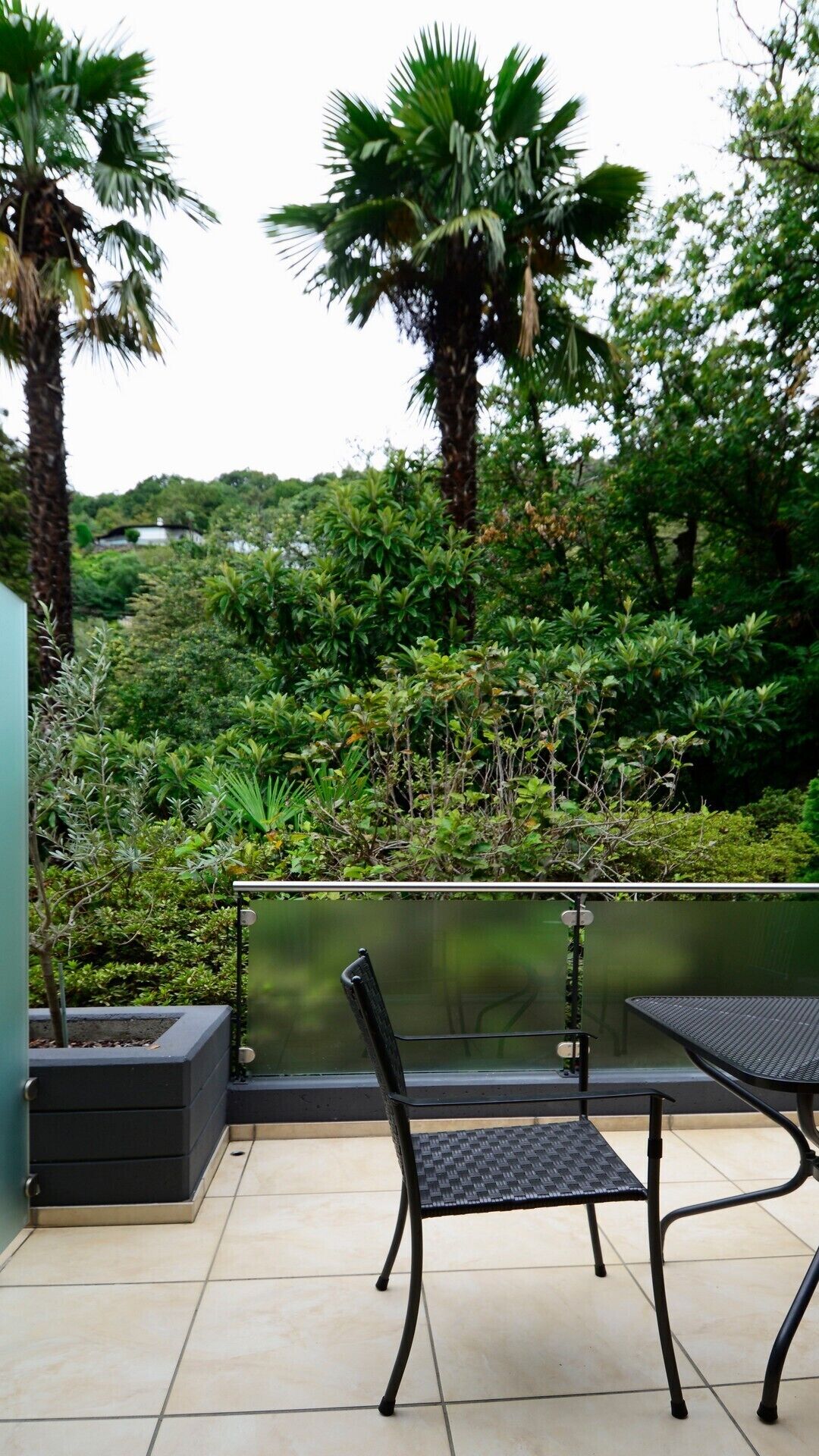 An outdoor patio with two black metal chairs and a table, surrounded by lush greenery and a view of a forested area.