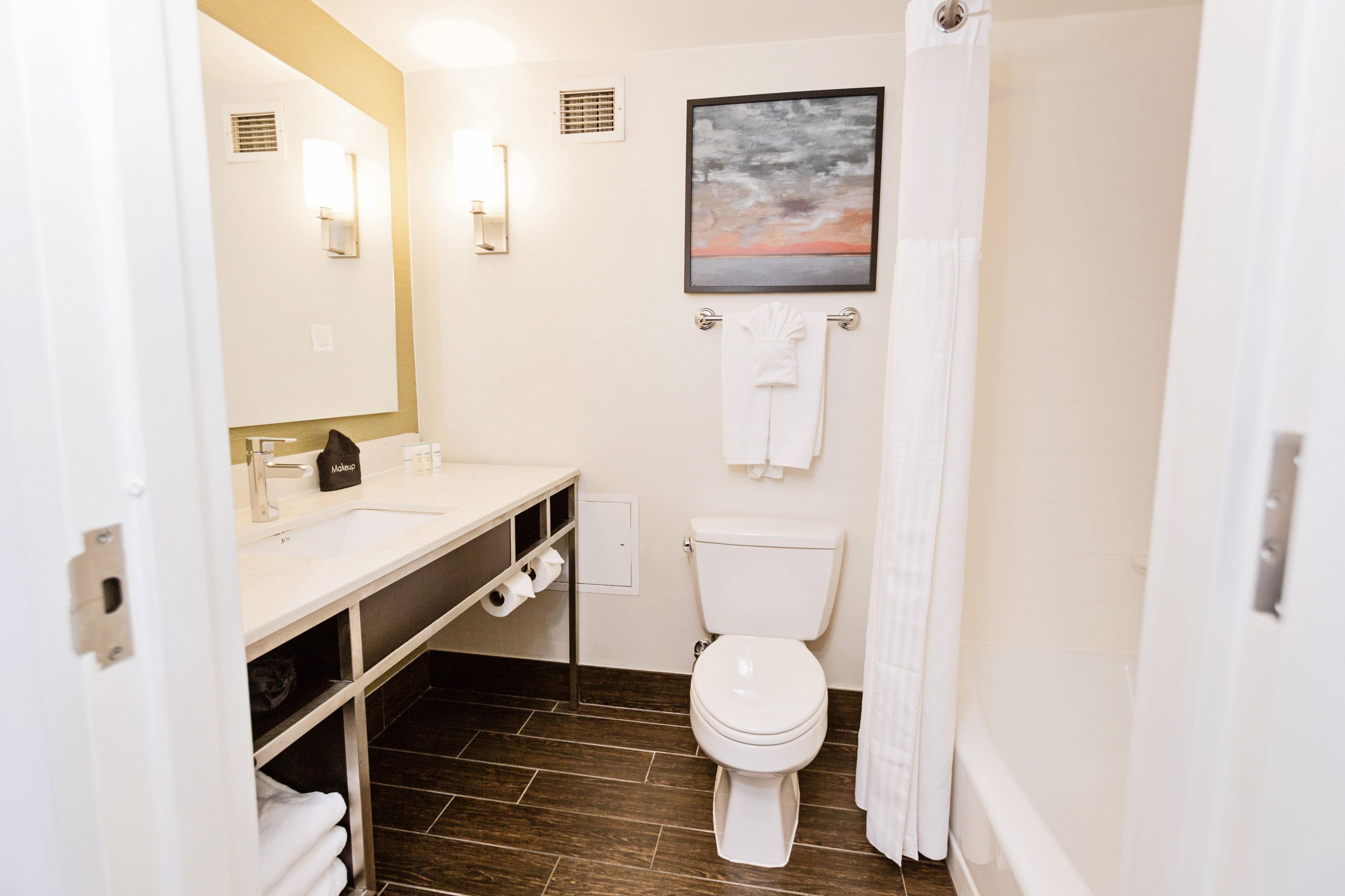 A bathroom with a white toilet, a sink with a mirror, a towel rack, and a painting on the wall.