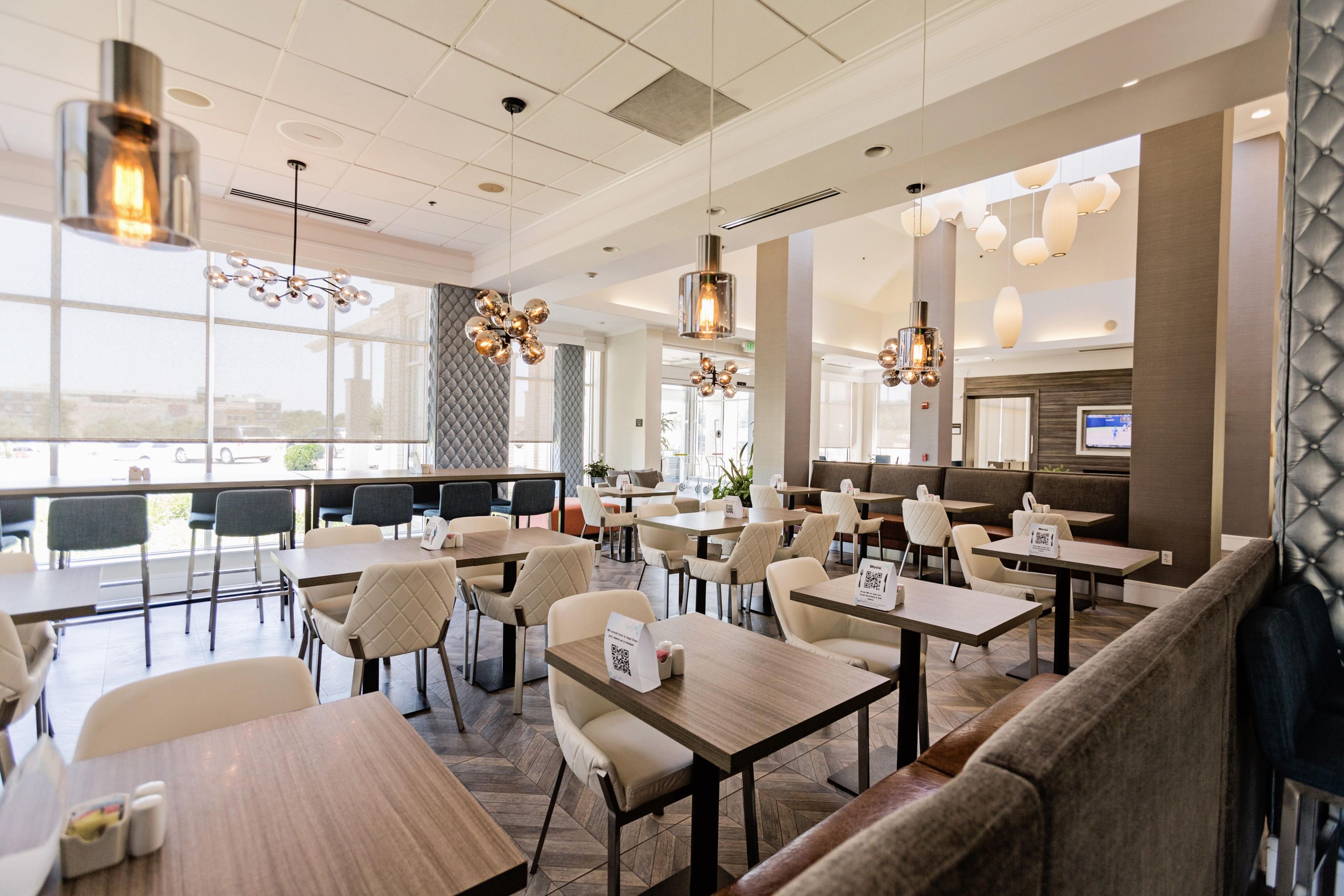 A modern dining area with multiple tables and chairs, a seating area with cushioned benches, and a large window offering a view.