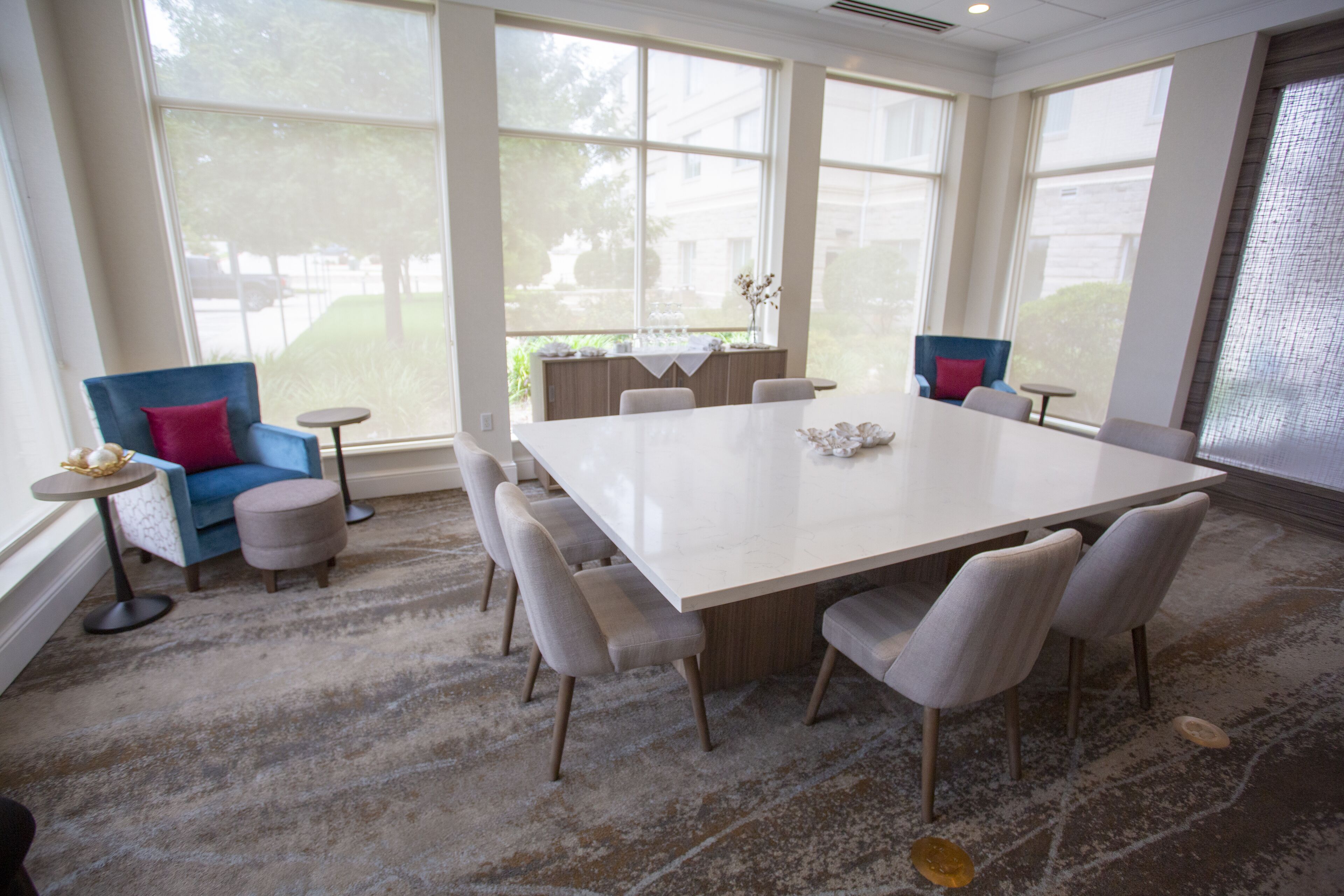 A spacious room with a large white table surrounded by chairs, large windows, and a view of the outdoors.