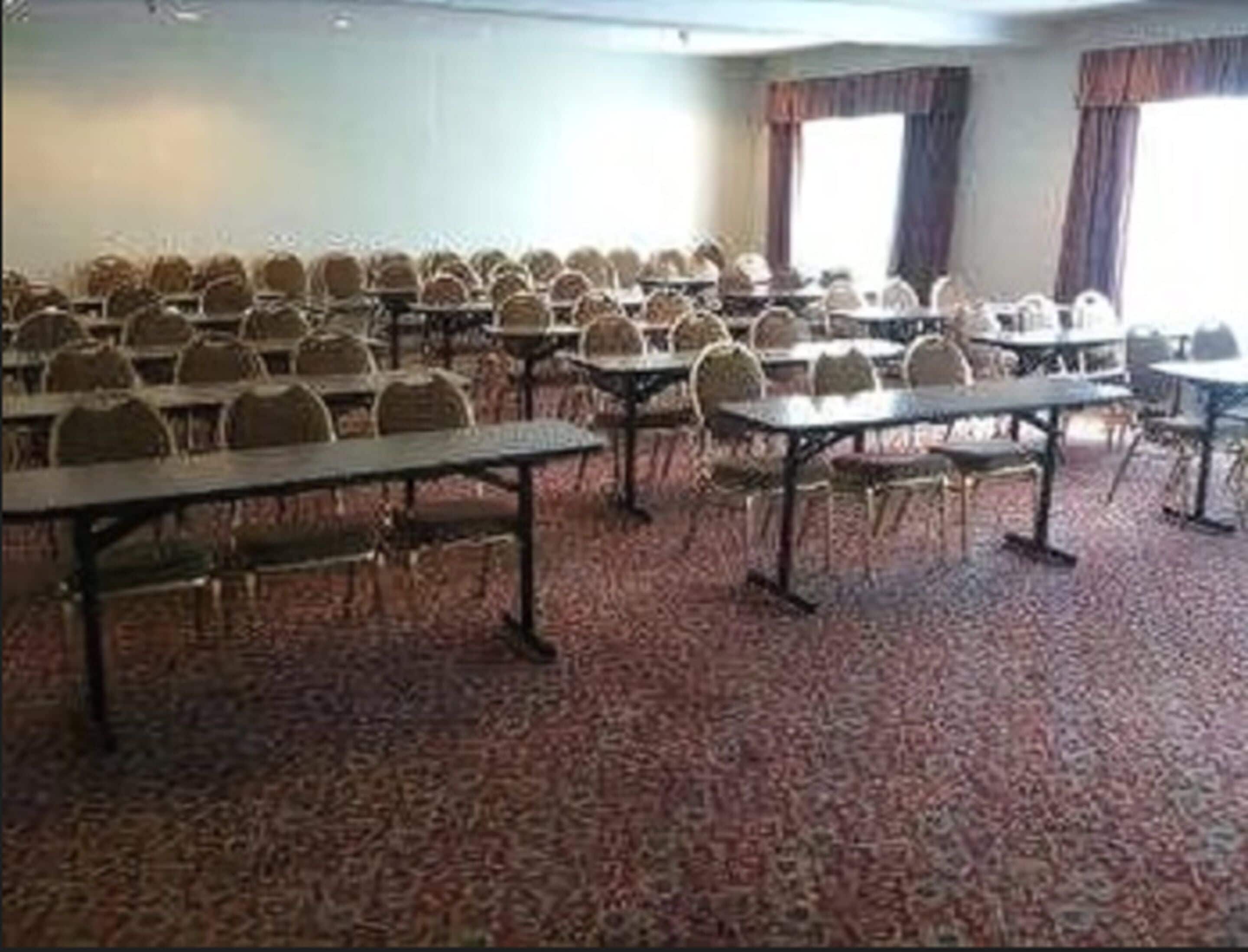 A conference room with rows of tables and chairs, carpeted flooring, and large windows with curtains.