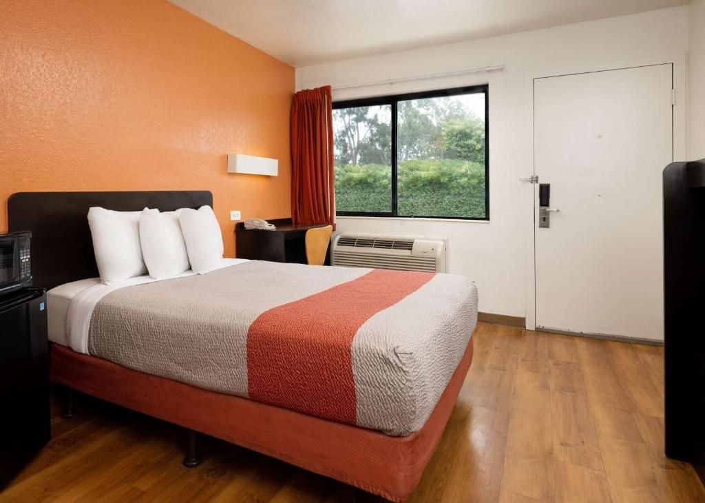 A hotel room with a bed, a nightstand, a desk, a window with a view, and a door.