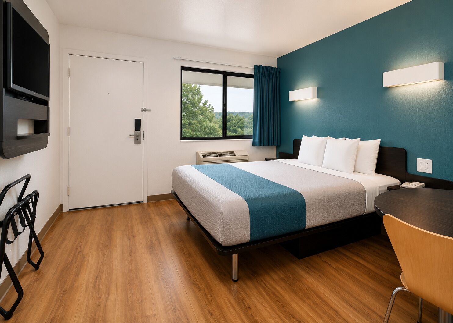 A hotel room with a bed, a desk, a chair, a TV, and a window with a view of trees.