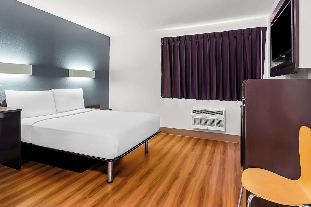 A hotel room with a bed, a nightstand, a chair, a window with curtains, and a wall-mounted air conditioning unit.