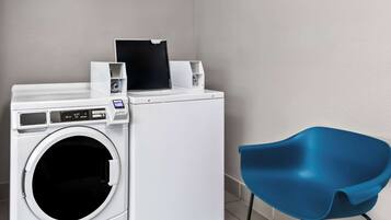 A compact, modern laundry room with a front-loading washer and dryer, a blue chair, and a small table with a black object on top.
