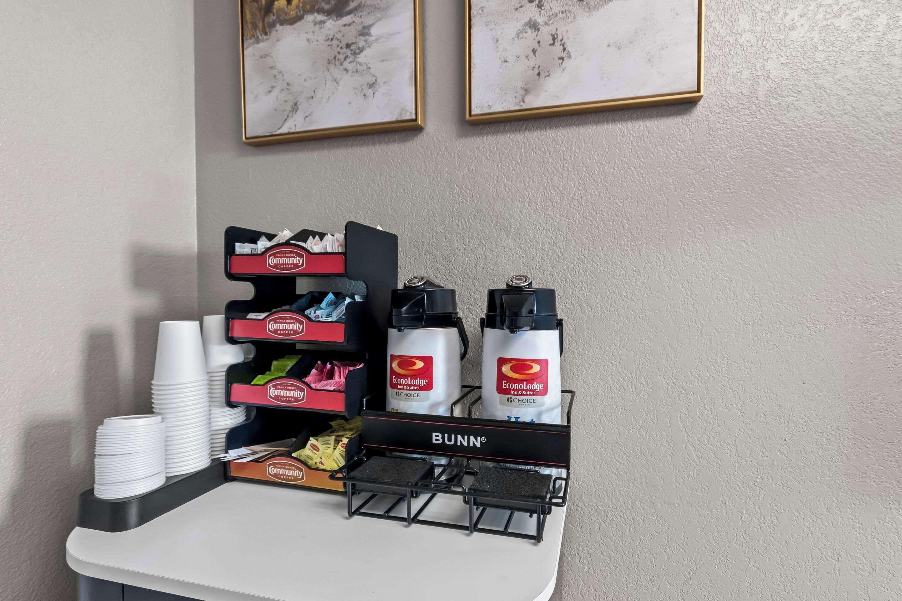 A hotel reception area with a coffee station featuring branded dispensers, a stack of white cups, and a Bunn coffee machine.