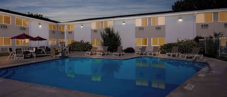 A swimming pool with lounge chairs and umbrellas, surrounded by a building with multiple windows and a fenced area.