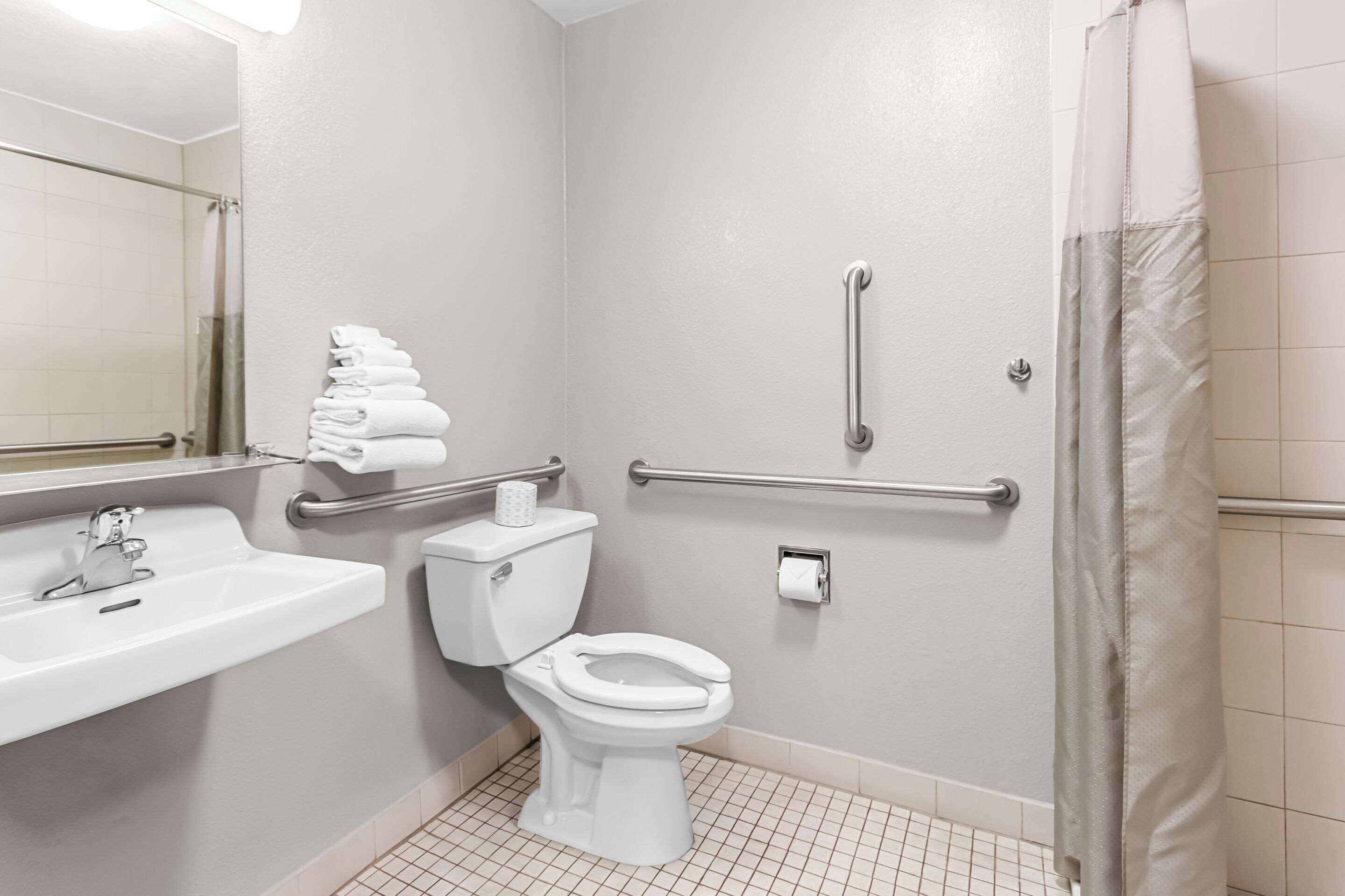 Une salle de bain avec un lavabo, des toilettes et une douche, le tout de couleur blanche. Des barres d’appui sont fixées aux murs, et un porte-serviettes contient des serviettes.
